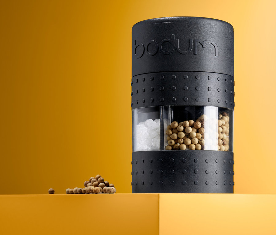 Černý mlýnek na sůl a pepř Bodum „Bistro“ stojí na žluto-oranžovém povrchu, vedle něj je malá hromádka kuliček pepře.