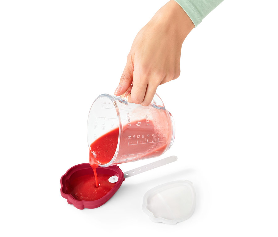 Člověk nalévá červenou tekutinu z odměrky do červené formičky na nanuky z 4 ks sady.