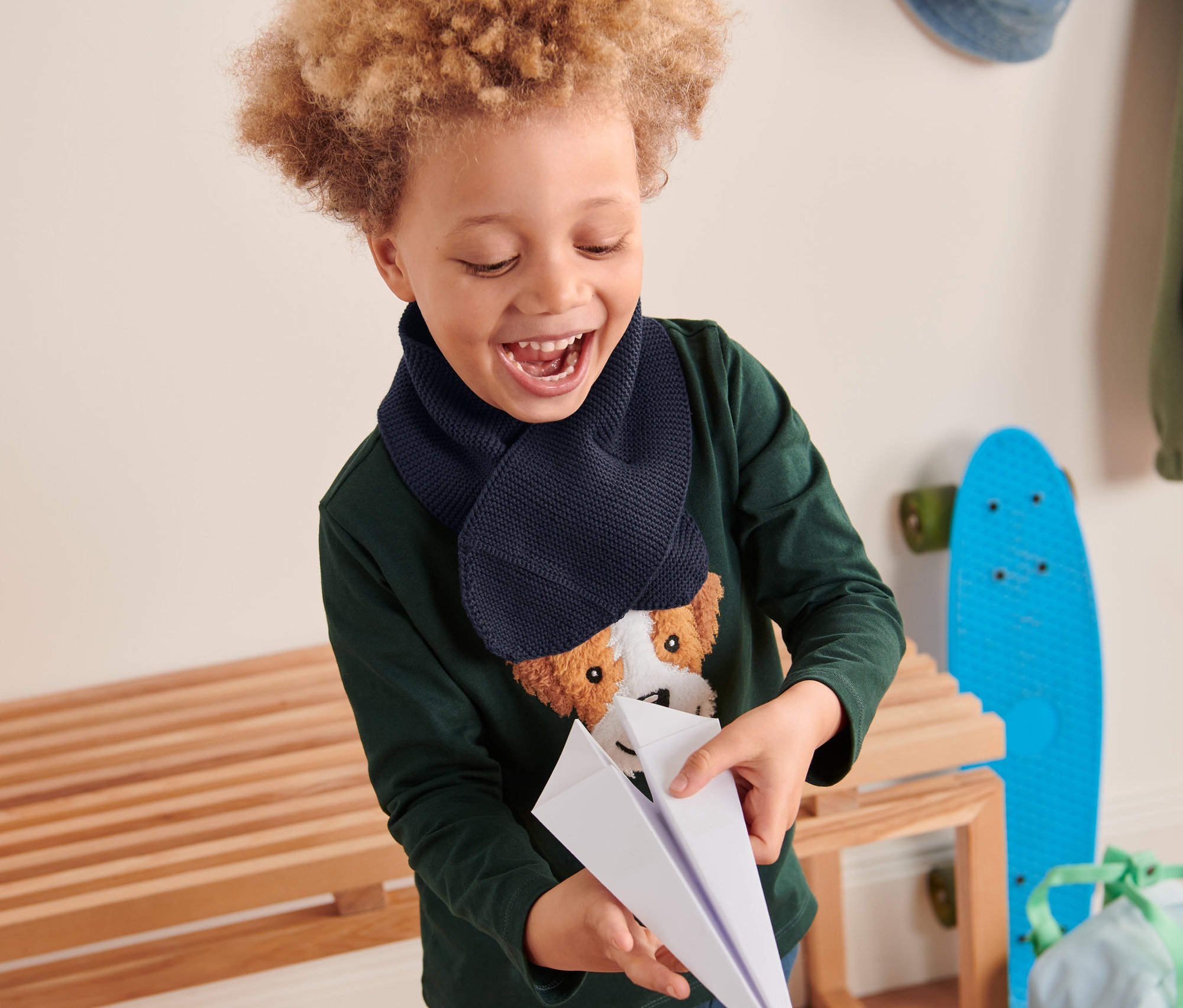 Chlapec se kudrnatými vlasy se směje a drží papírové letadlo. Má na sobě tmavě modrou pletenou šálu.
