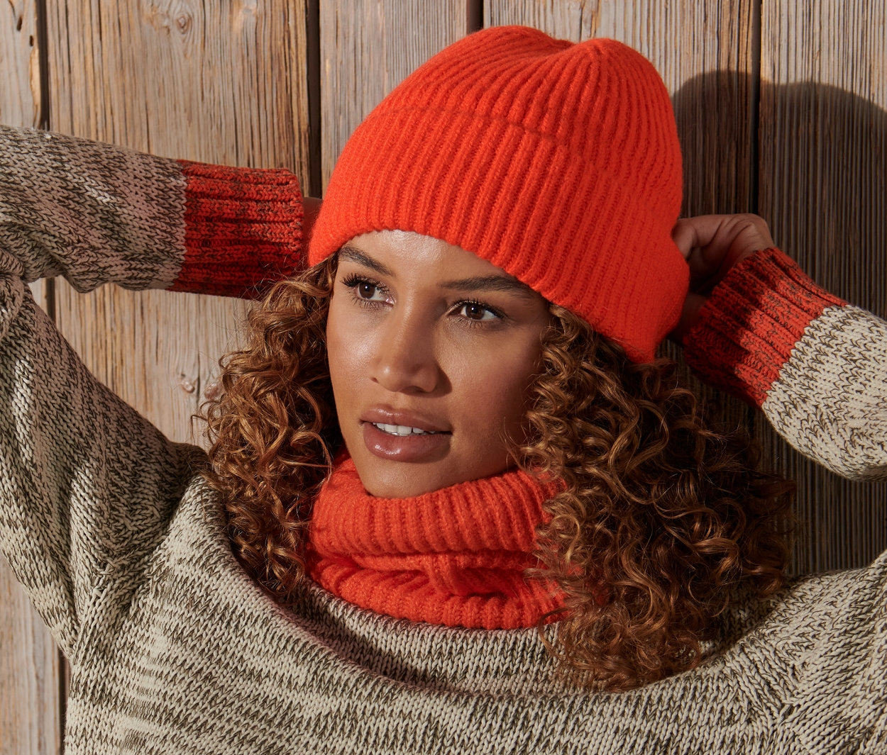 Žena s kudrnatými vlasy pózuje s rukama za hlavou a má na sobě oranžovou pletenou čepici.