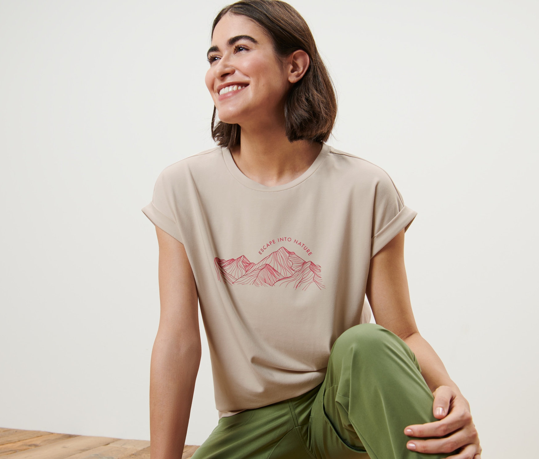 Žena sedí na dřevěné podlaze a má na sobě béžové tričko s potiskem hory.