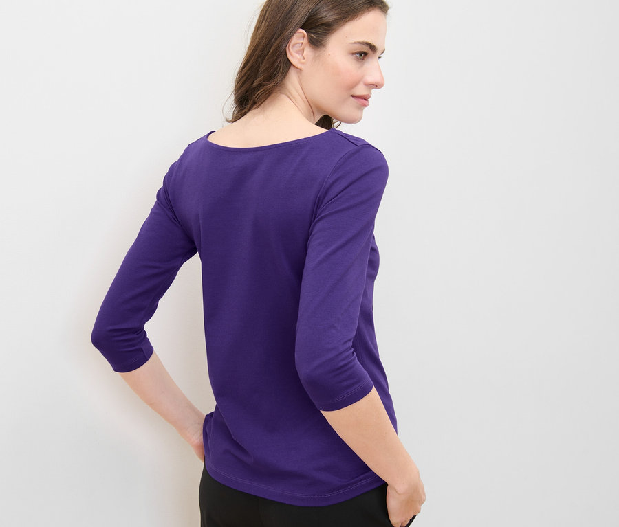 Žena pózuje v fialovém tričku s tříčtvrtečním rukávem.