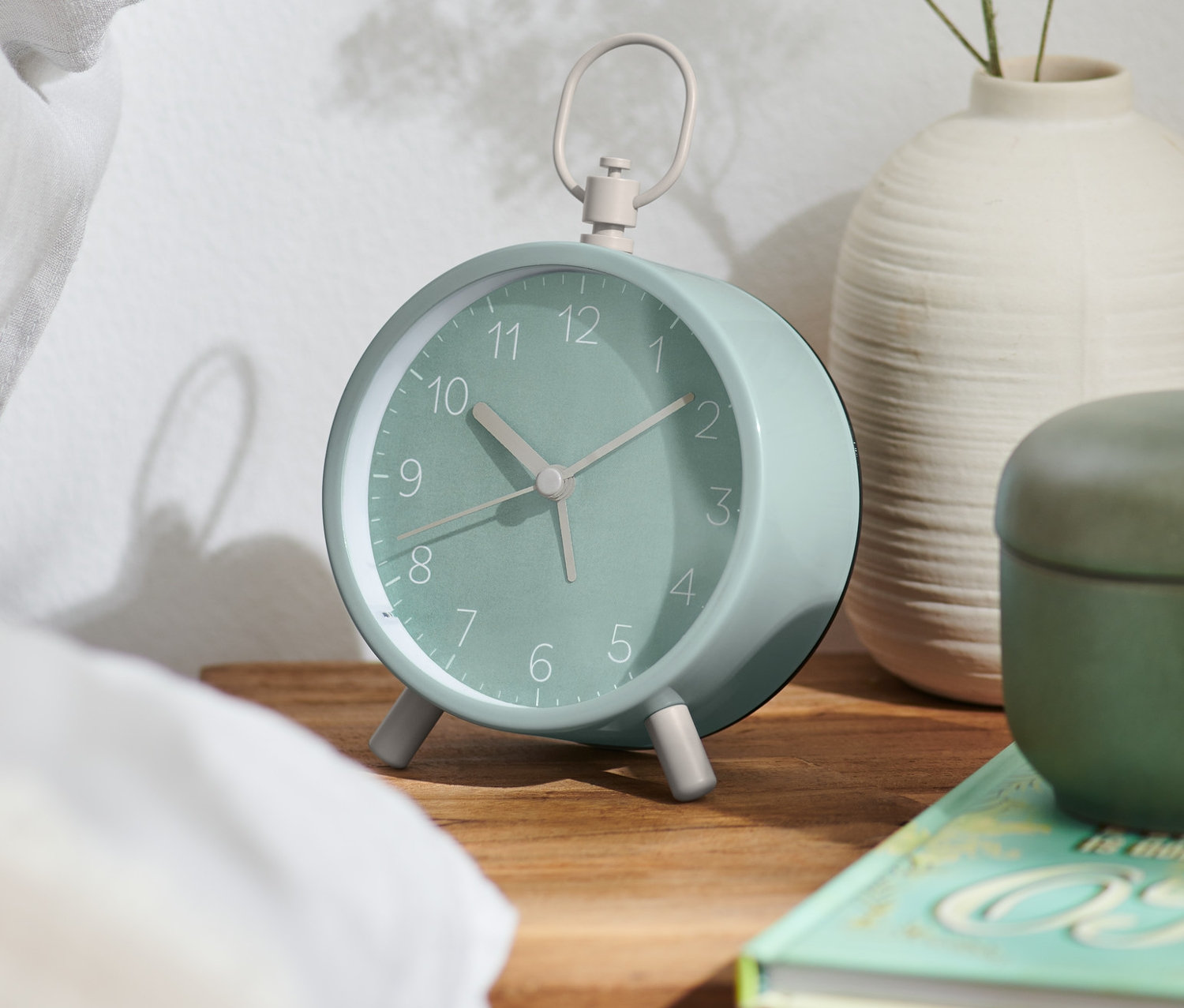 Zelený designový analogový budík stojí na dřevěném nočním stolku vedle postele.