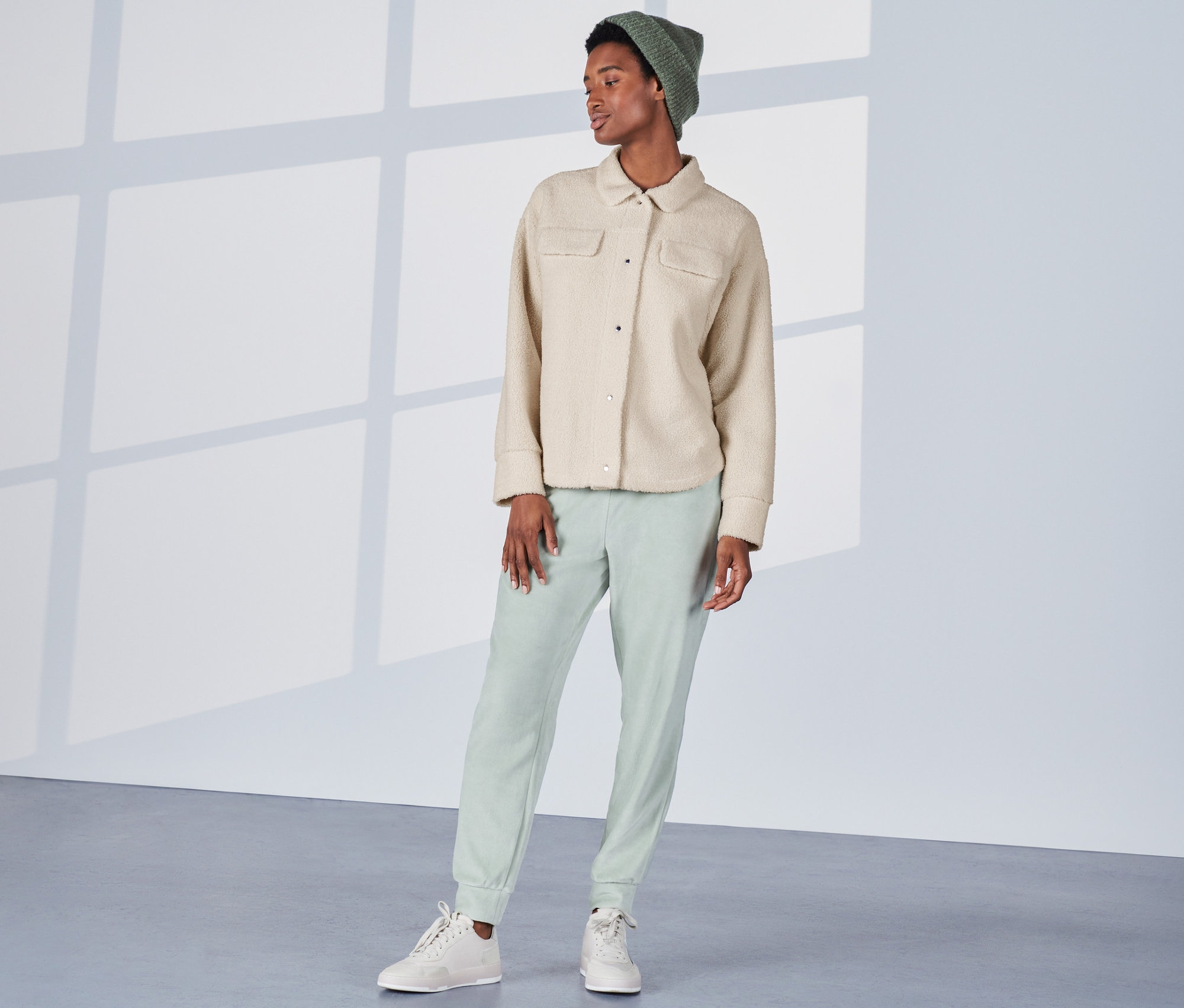 Žena stojí a má na sobě krémově bílou plyšovou svrchní košili, zelené kalhoty a čepici.