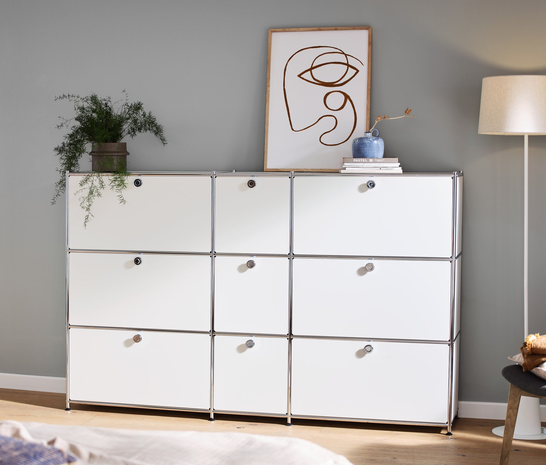 V pokoji stojí bílá komoda CN3 s 9 výklopnými dvířky, na níž je dekorace. Vlevo je konzolový stolek a stojací lampa s textilním stínítkem.