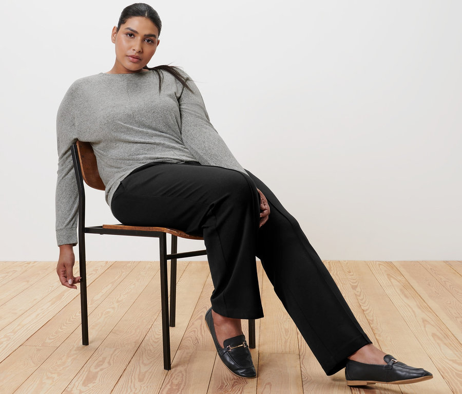 Žena sedí na židli a má na sobě černé žerzejové kalhoty s puky, triko s netopýřími rukávy a kožené mokasíny.