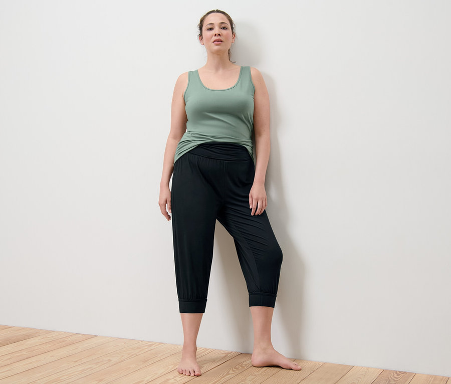 Žena stojí a má na sobě zelené tílko a černé 3/4 kalhoty na jógu.