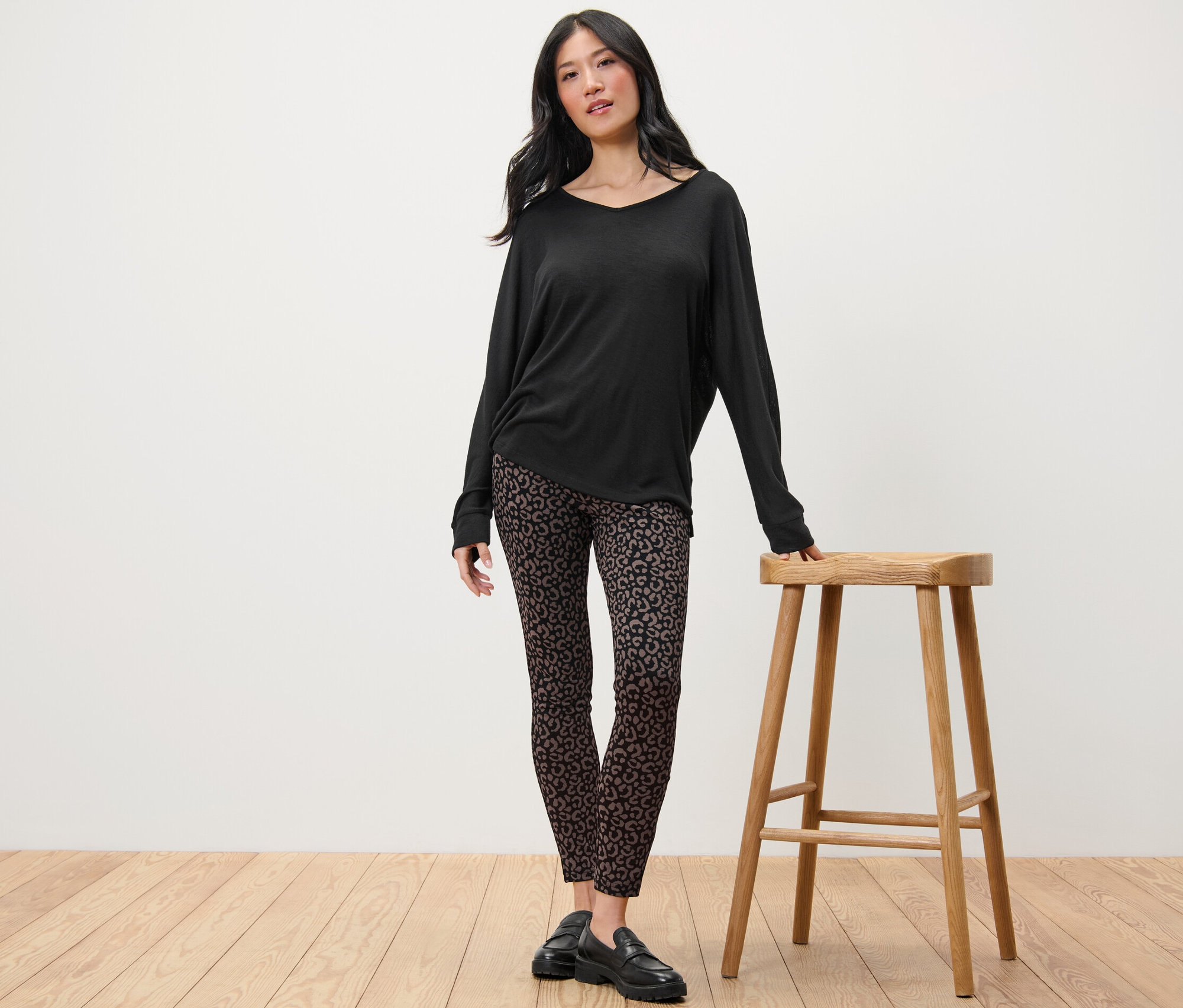 Žena stojí vedle dřevěné stoličky a má na sobě černé tričko s dlouhým rukávem a pevné legíny s potiskem.