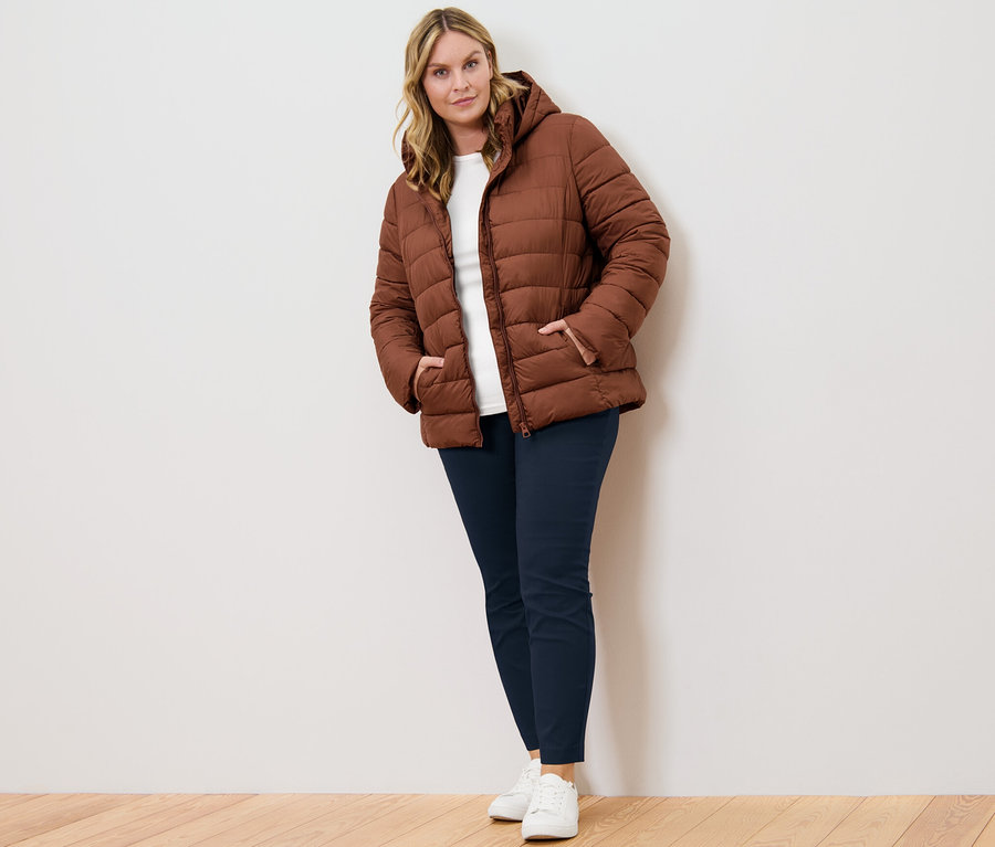 Žena pózuje v koňakové prošívané bundě s kapucí, tmavě modrých strečových kalhotách a bílém triku s 3/4 rukávem.