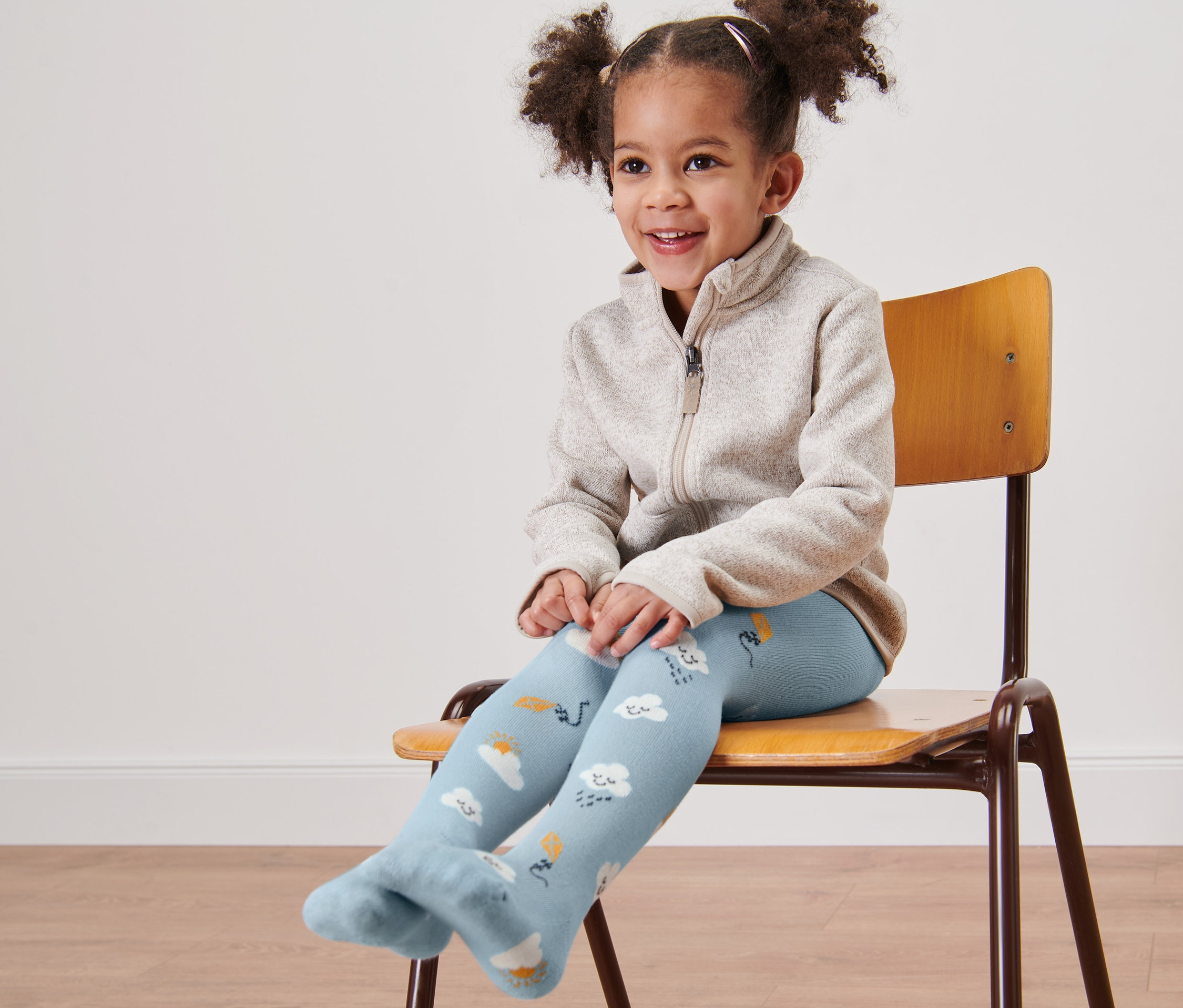 Usmívající se holčička sedí na židli a má na sobě modré punčocháče s obláčky.