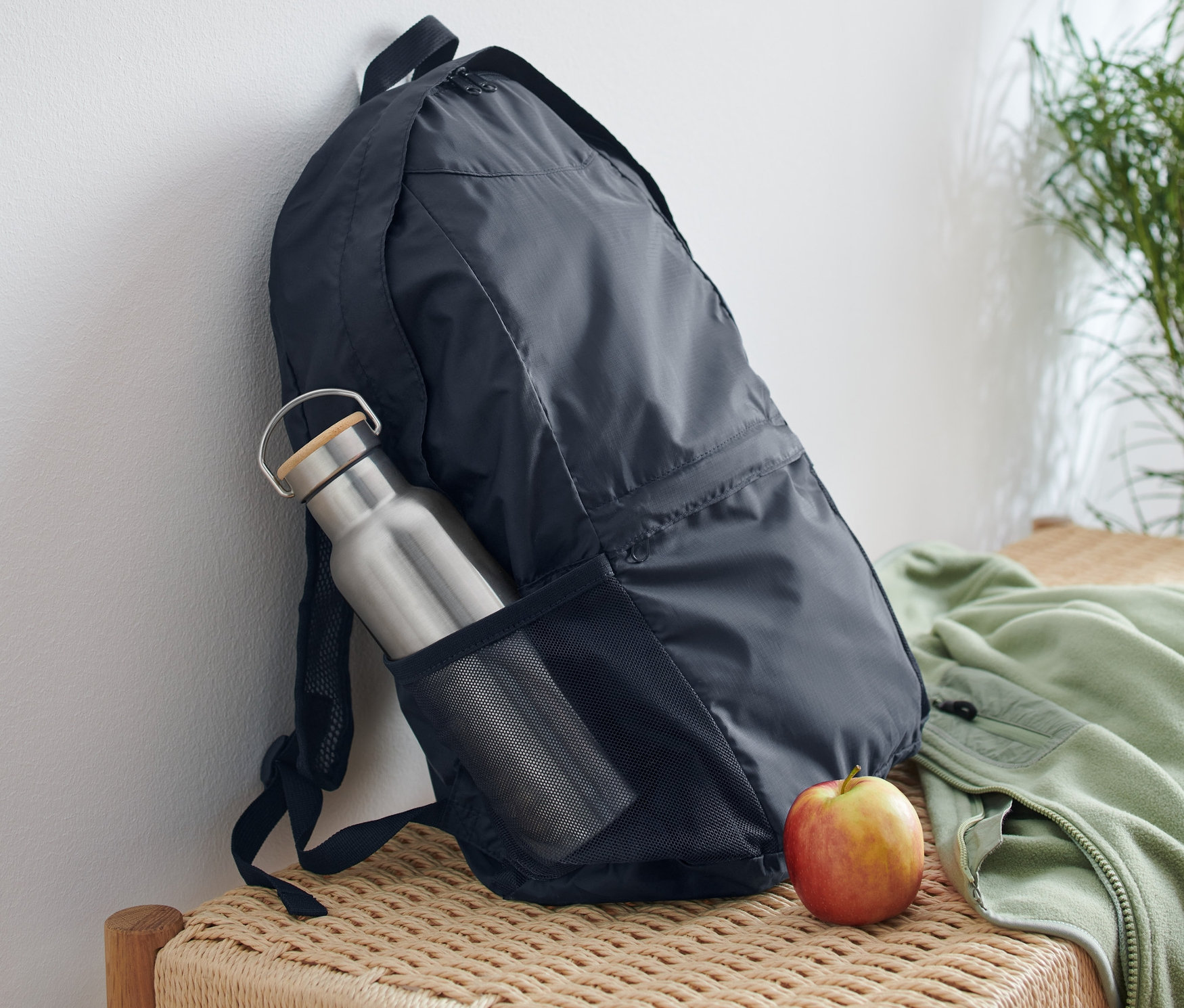 Skládací batoh opřený o bílou zeď s lahví na vodu v boční kapse a jablkem vedle něj na proutěné lavici.