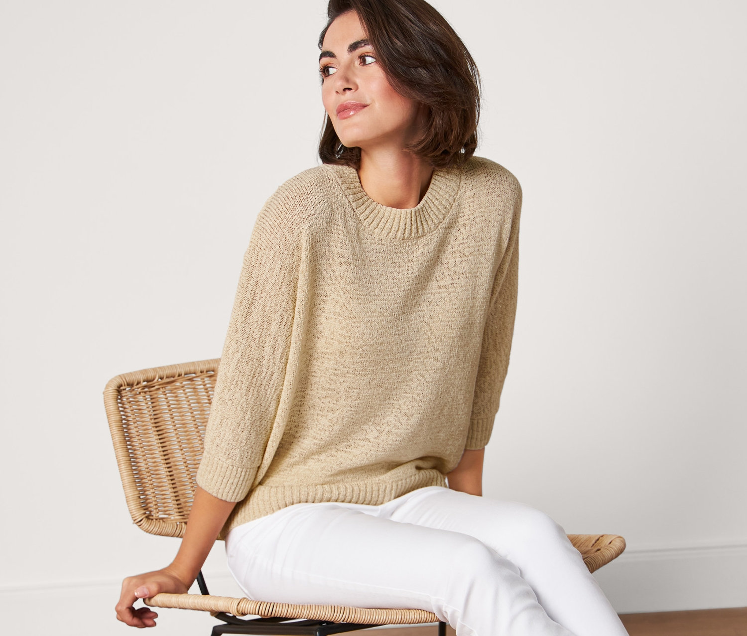 Žena sedí na židli a má na sobě letní pletený svetr z páskové příze, bílé kalhoty a tenisky.
