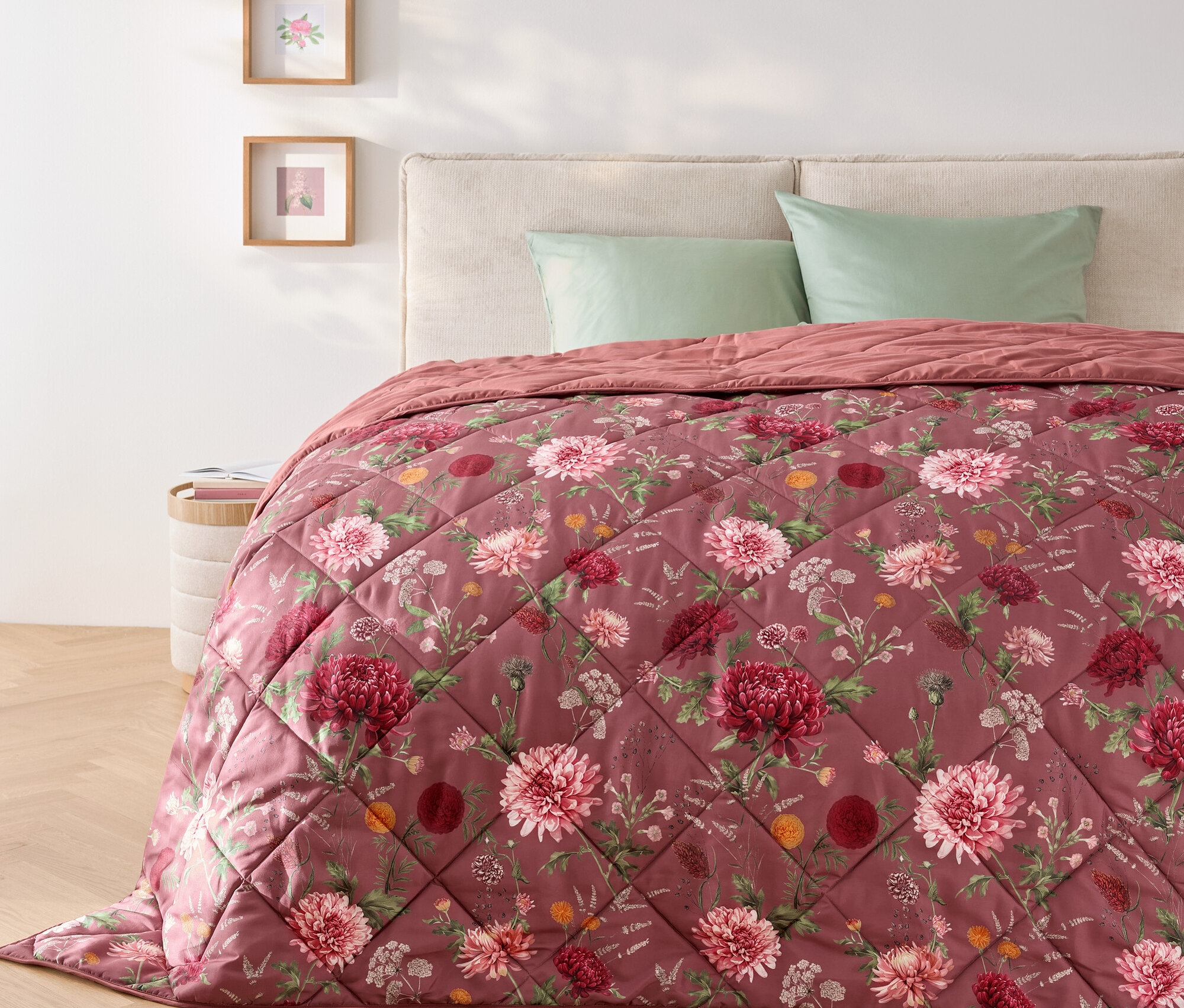 Na posteli leží prošívaná přikrývka s květinovým vzorem a dva zelené polštáře.