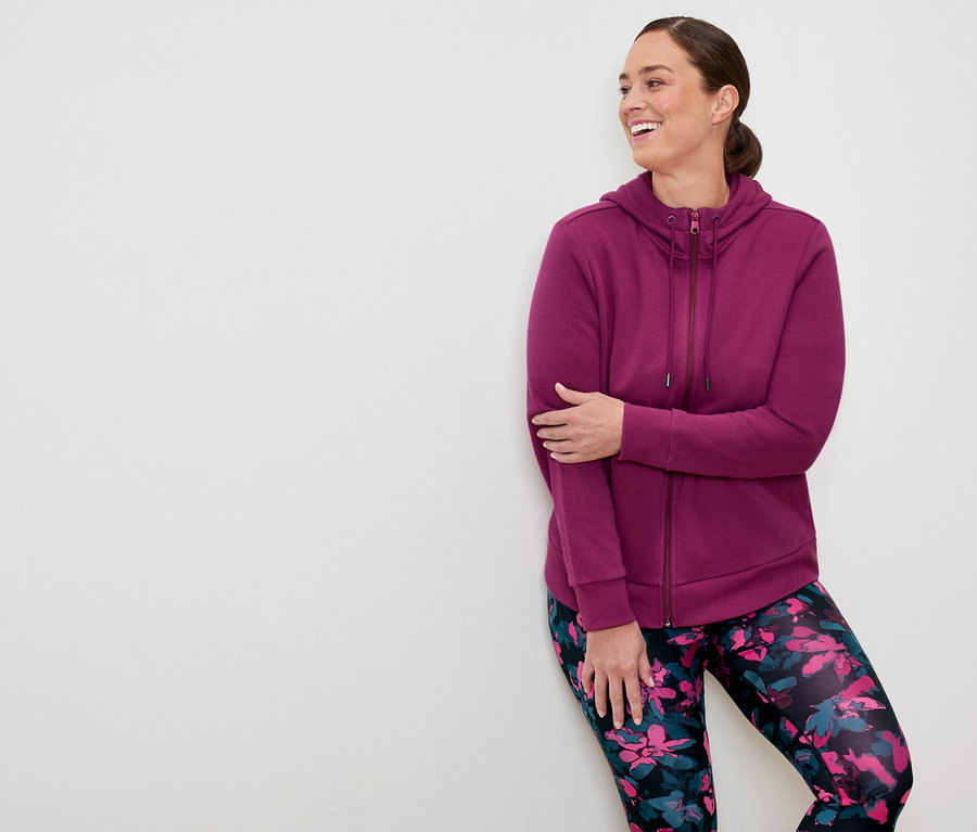 Žena v purpurové mikině na zip a květinových legínách se opírá o bílou zeď a usmívá se.