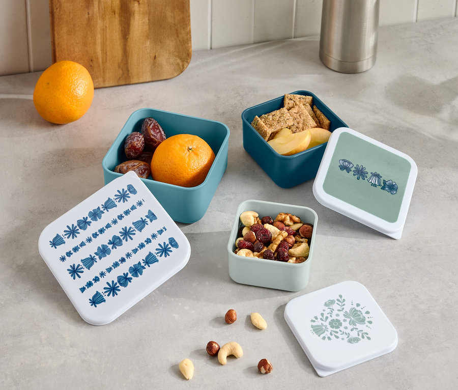 Na lince stojí termoska a dřevěné prkénko vedle čtyř malých otevřených krabiček s ovocem, krekry a ořechy a čtyř víček s různými vzory.