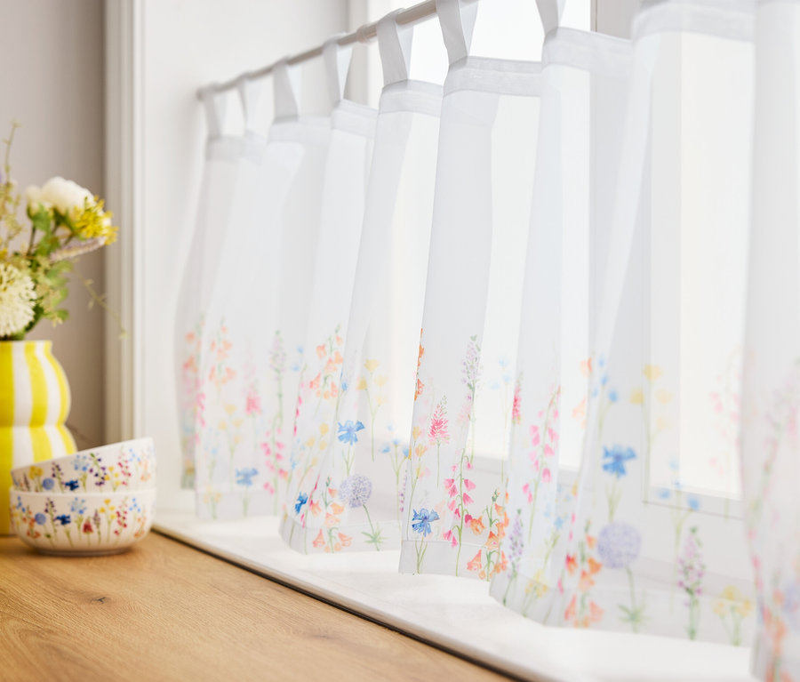 Květiny ve žluté váze a dvě misky na dřevěné desce před okny se záclonami s květinovým vzorem.