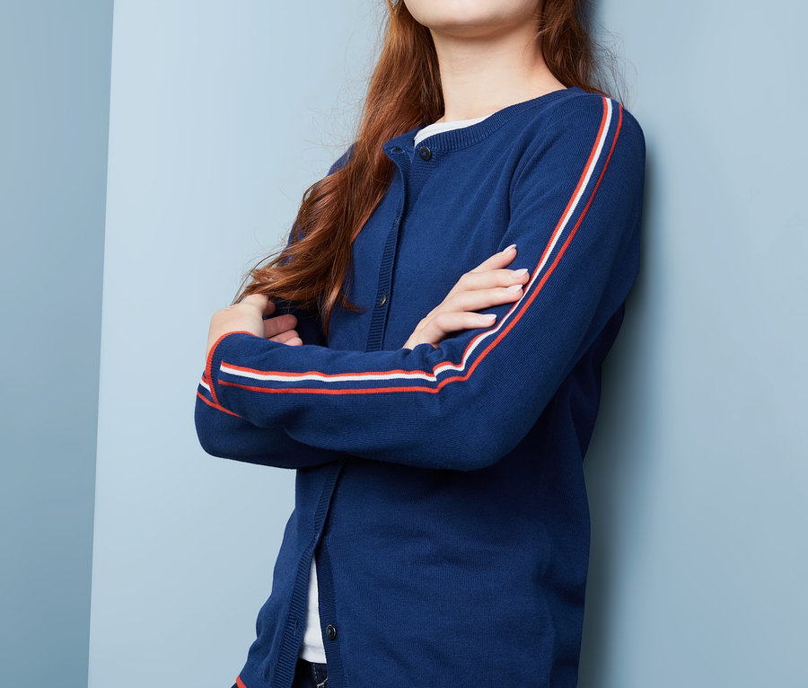 Dívka s hnědými vlasy pózuje v pleteném kabátku s modrými pruhy na rukávech.