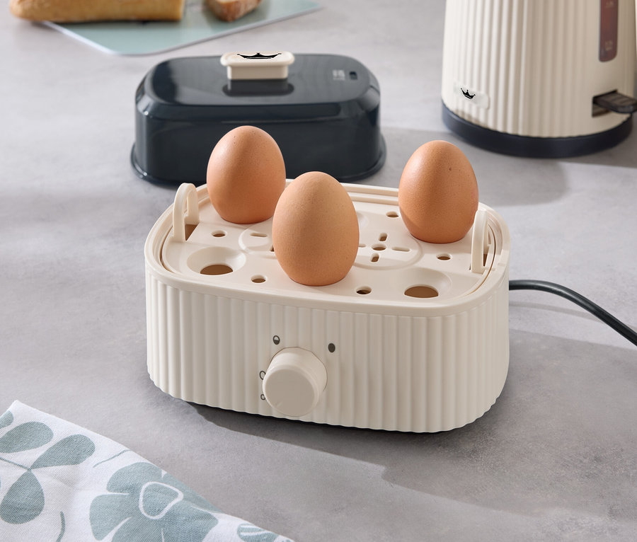 Tři vejce ve vařiči vajec na kuchyňské lince, v pozadí toustovač, rychlovarná konvice a chléb.