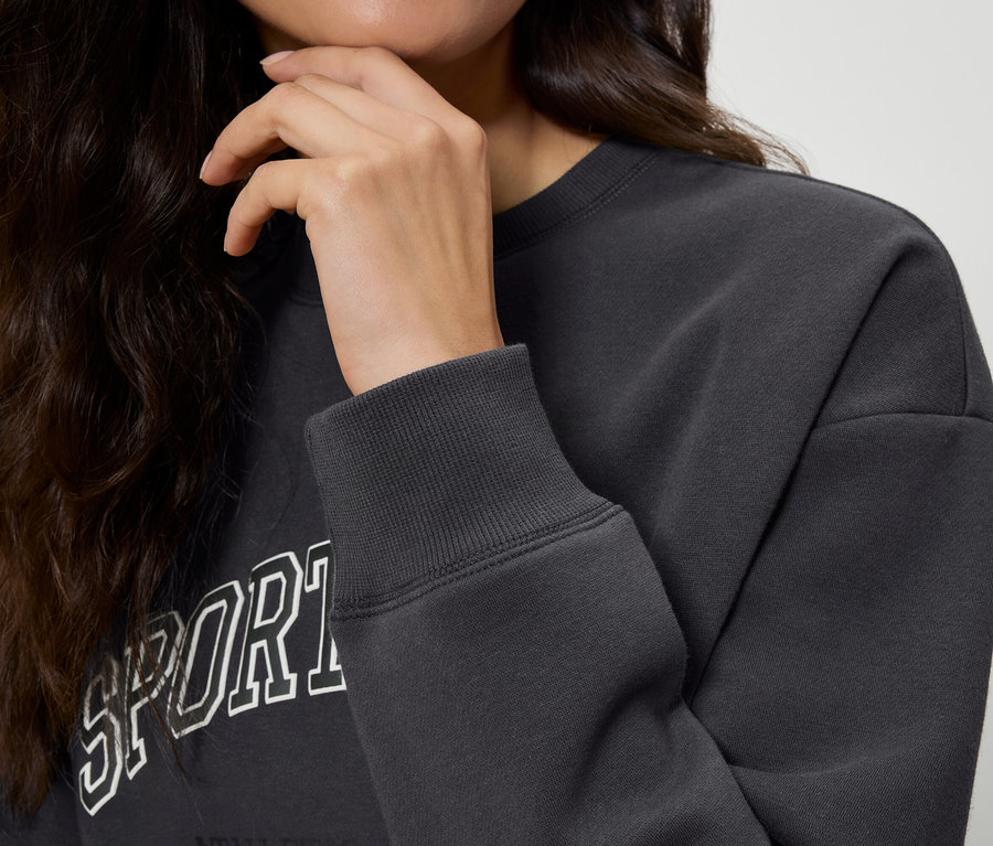 Částečný pohled na ženu s tmavými vlnitými vlasy, která se dotýká brady. Má na sobě tmavě šedou mikinu s nápisem Sport. Rukáv mikiny je v detailu.