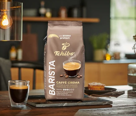 Balení BARISTA Caffè Crema – 1x 1 kg zrnkové kávy stojí na stole vedle sklenice kávy.