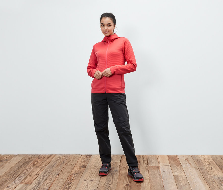 Žena pózuje v korálové mikině s kapucí, černých kalhotách a botách na dřevěné podlaze.