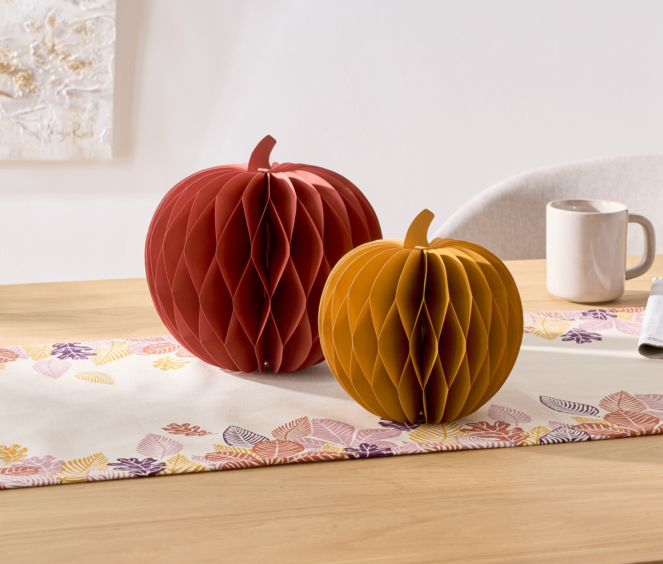 Na stole leží dva dekorativní papírové dýně, červená a žlutá. Vedle nich je hrnek a ubrousek.