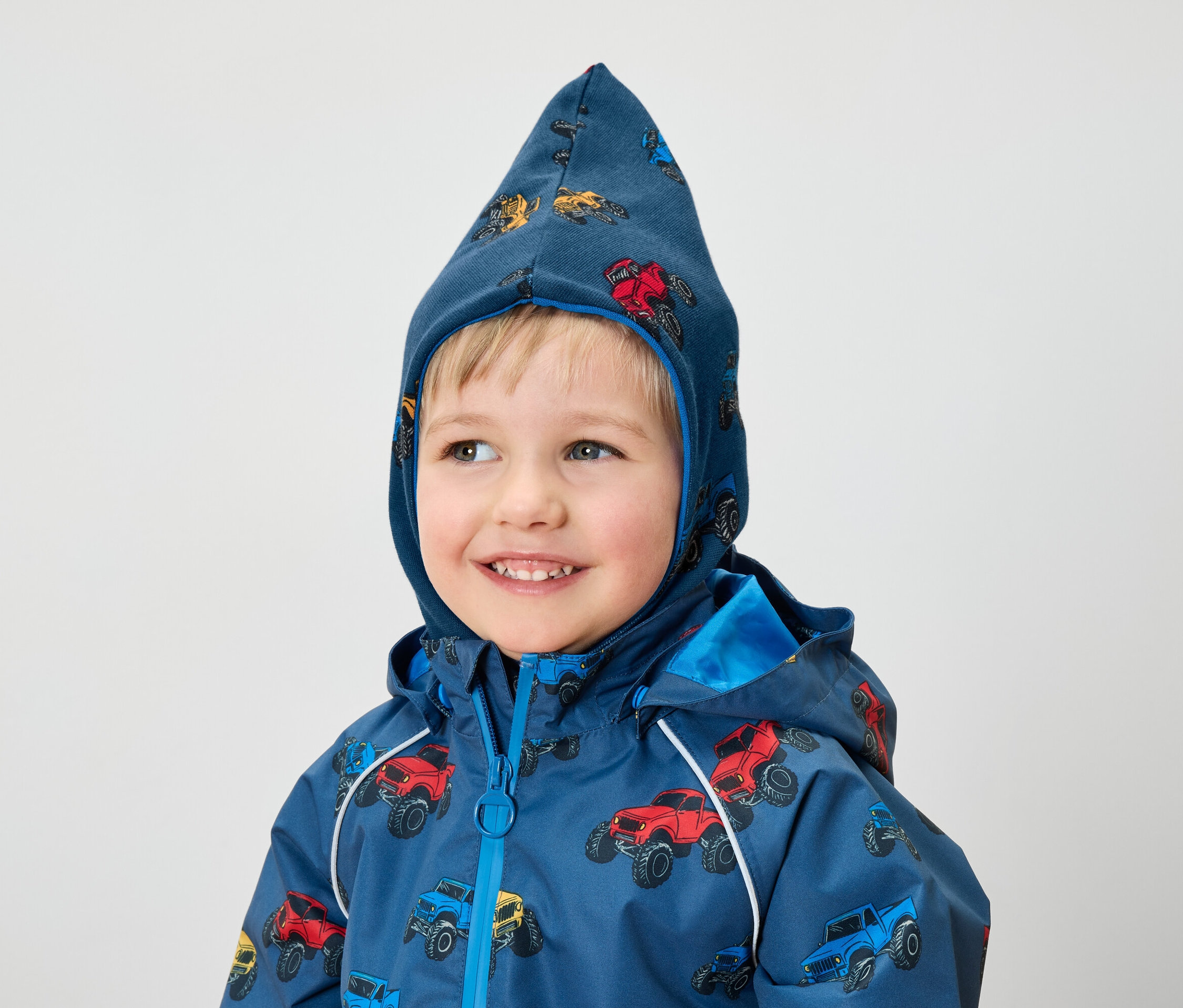 Usmívající se chlapec má na sobě modrou bundu a kapuci s potiskem monster trucků.