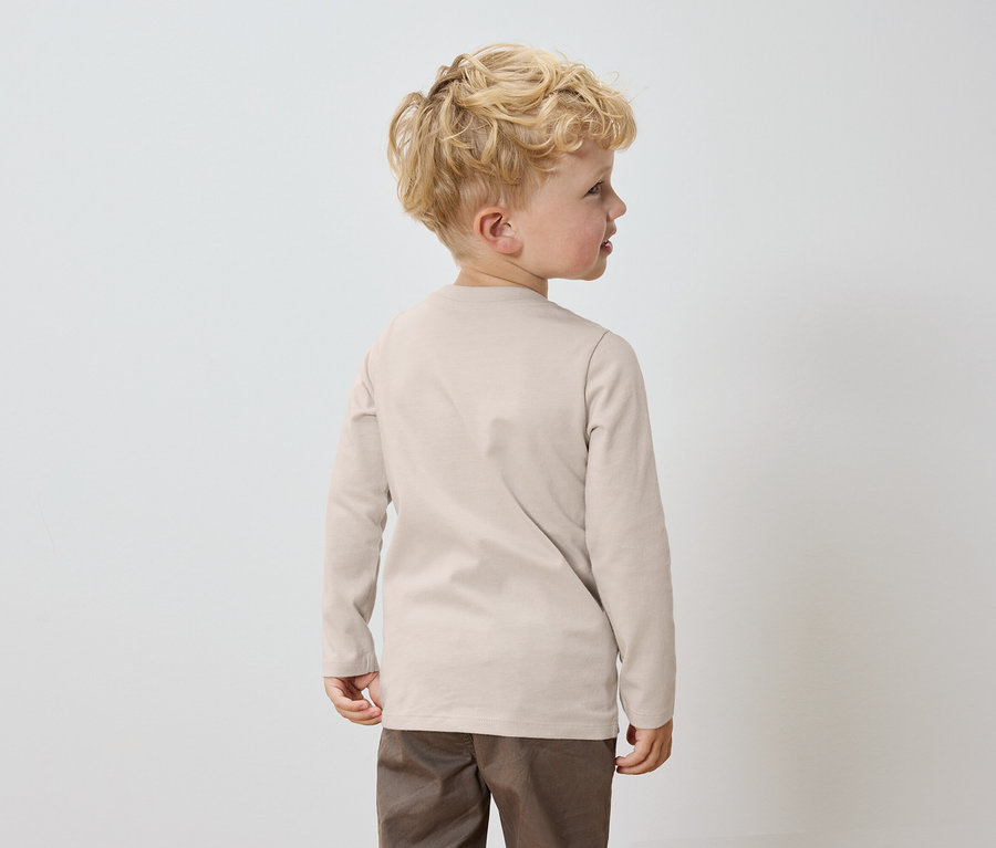 Chlapec s blonďatými vlasy stojí zády k fotoaparátu, oblečený v béžovém tričku s dlouhým rukávem a hnědých kalhotách.