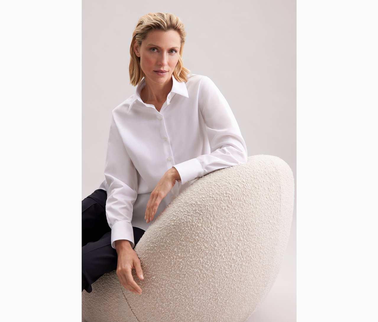 Žena sedí na křesle a má na sobě bílou dlouhou halenku oversize Seidensticker.