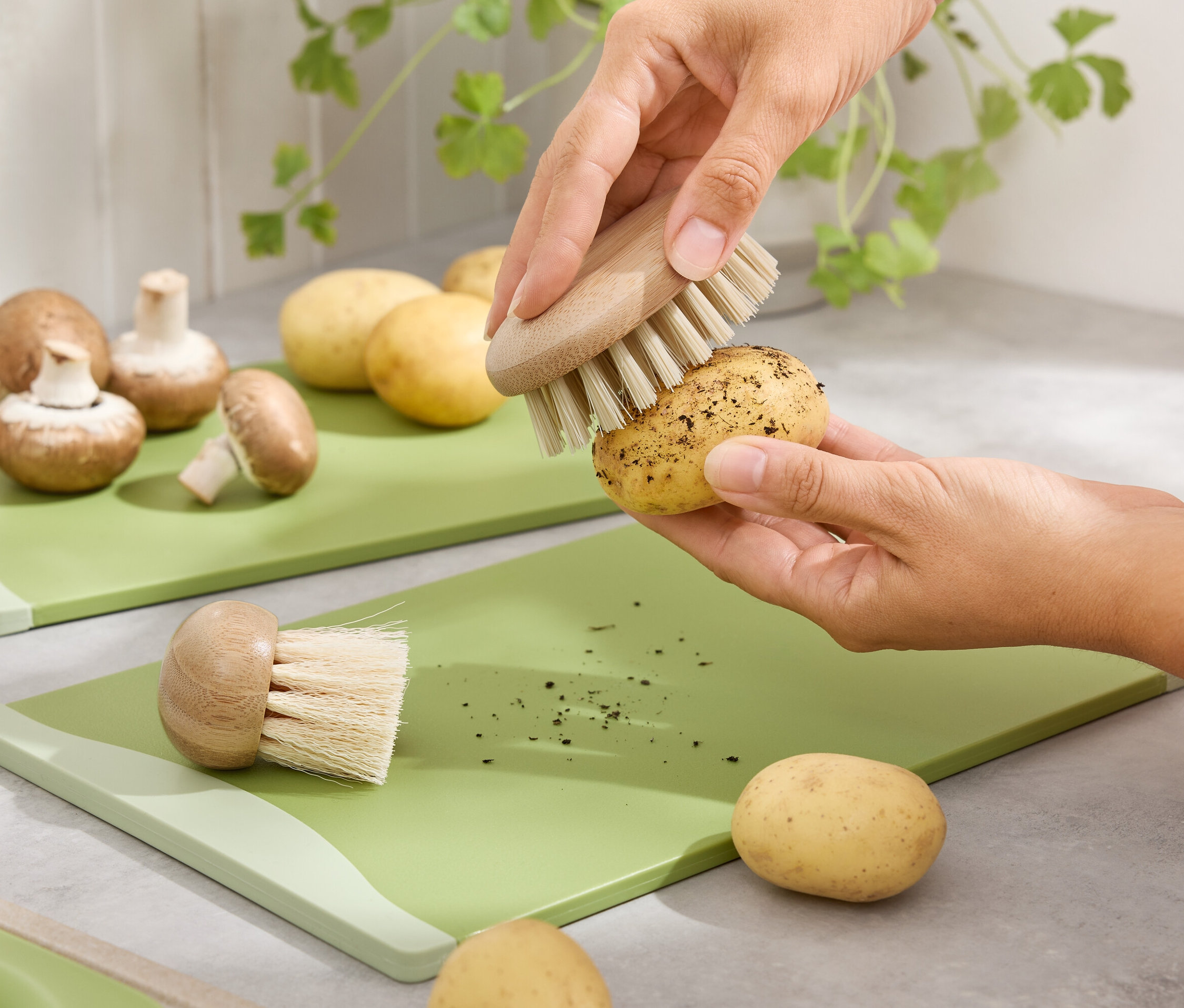 Ruce čistí kartáčem bramboru na zeleném prkénku. Hnědé žampiony a brambory jsou na pozadí.