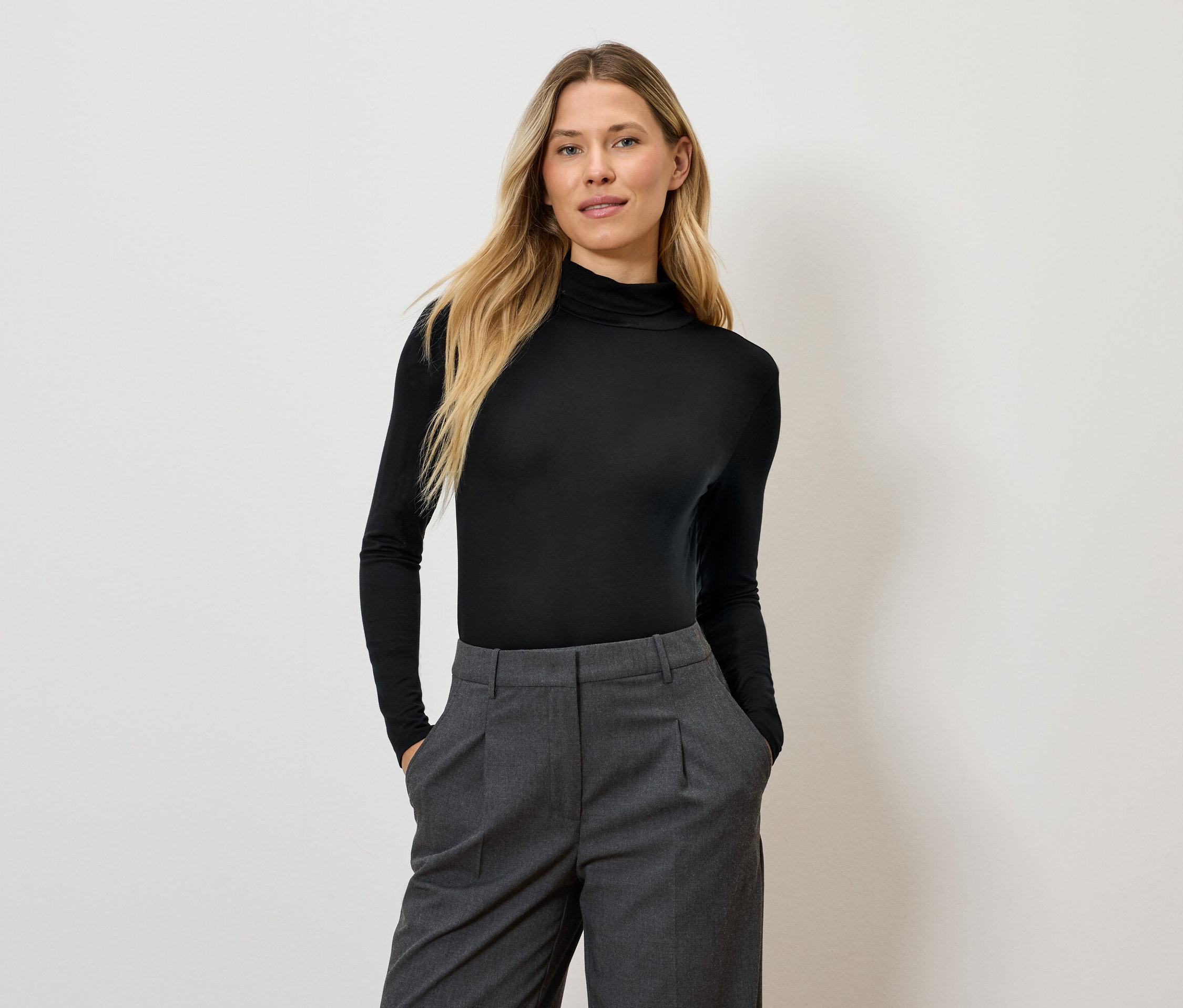 Žena s dlouhými blond vlasy pózuje v černém roláku a šedých kalhotách.