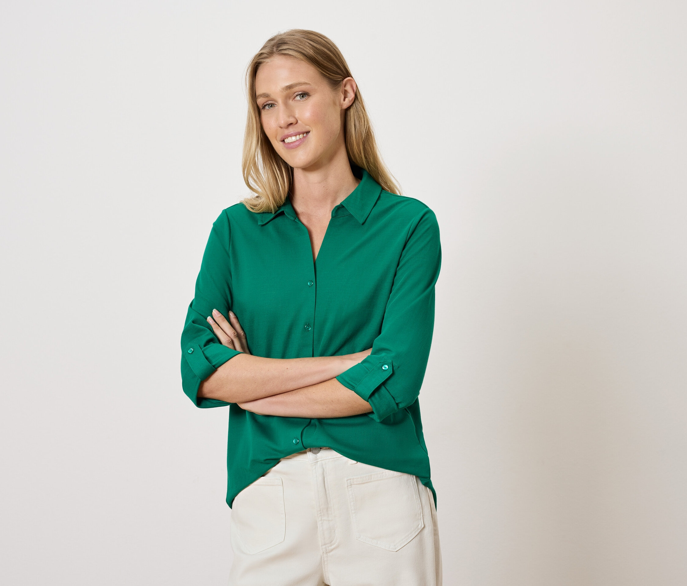 Žena se zkříženýma rukama pózuje v zelené košili a bílých kalhotách.