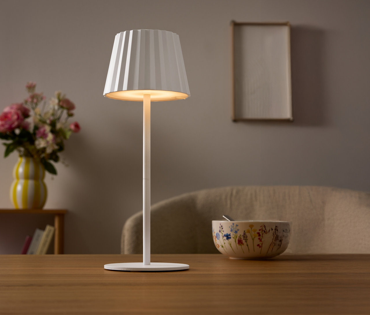 Na stole stojí bílá lampa s plisovaným stínidlem. V pozadí je váza s květinami a miska.