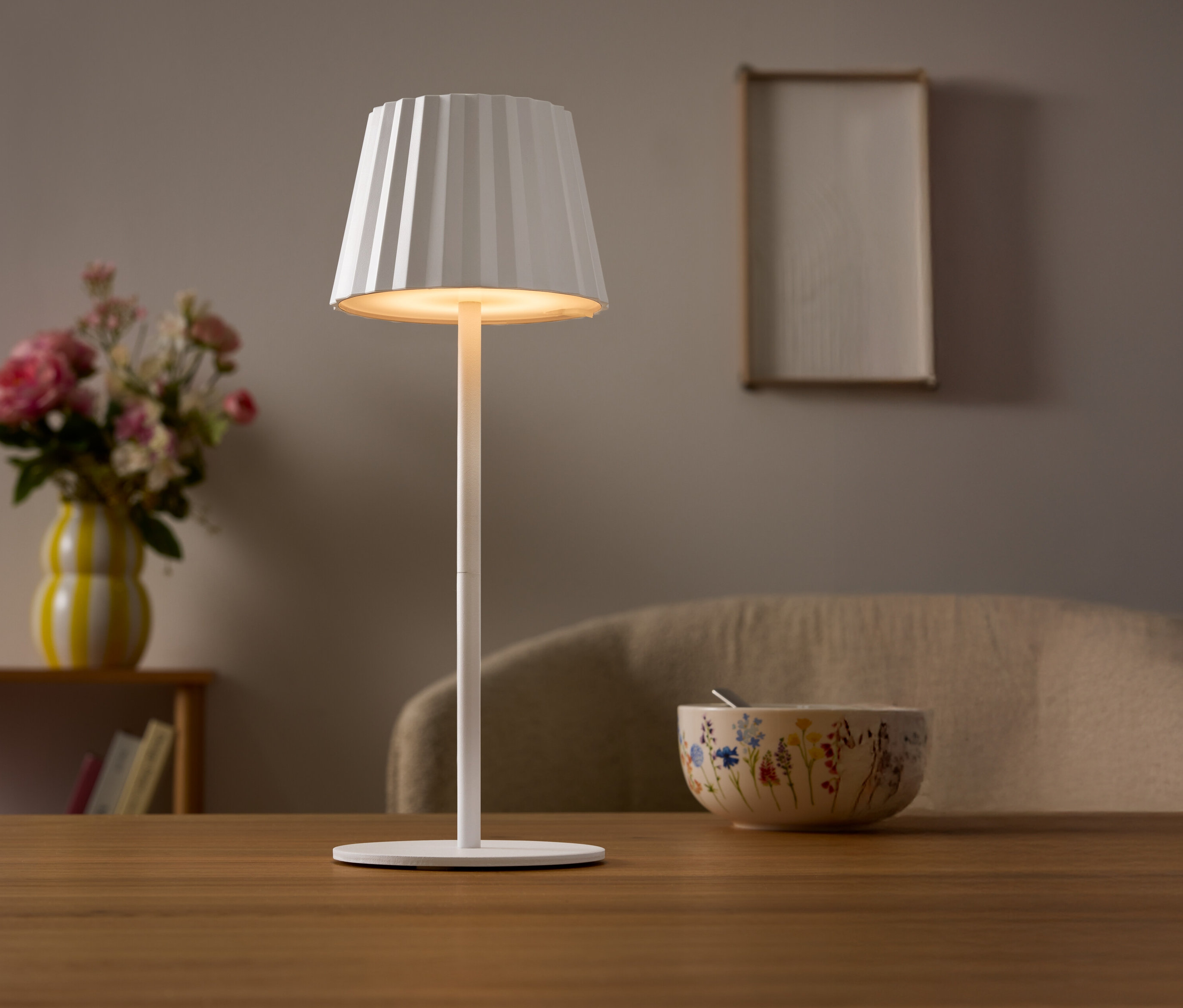 Na stole stojí bílá lampa s plisovaným stínidlem. V pozadí je váza s květinami a miska.