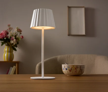 Na stole stojí bílá lampa s plisovaným stínidlem. V pozadí je váza s květinami a miska.