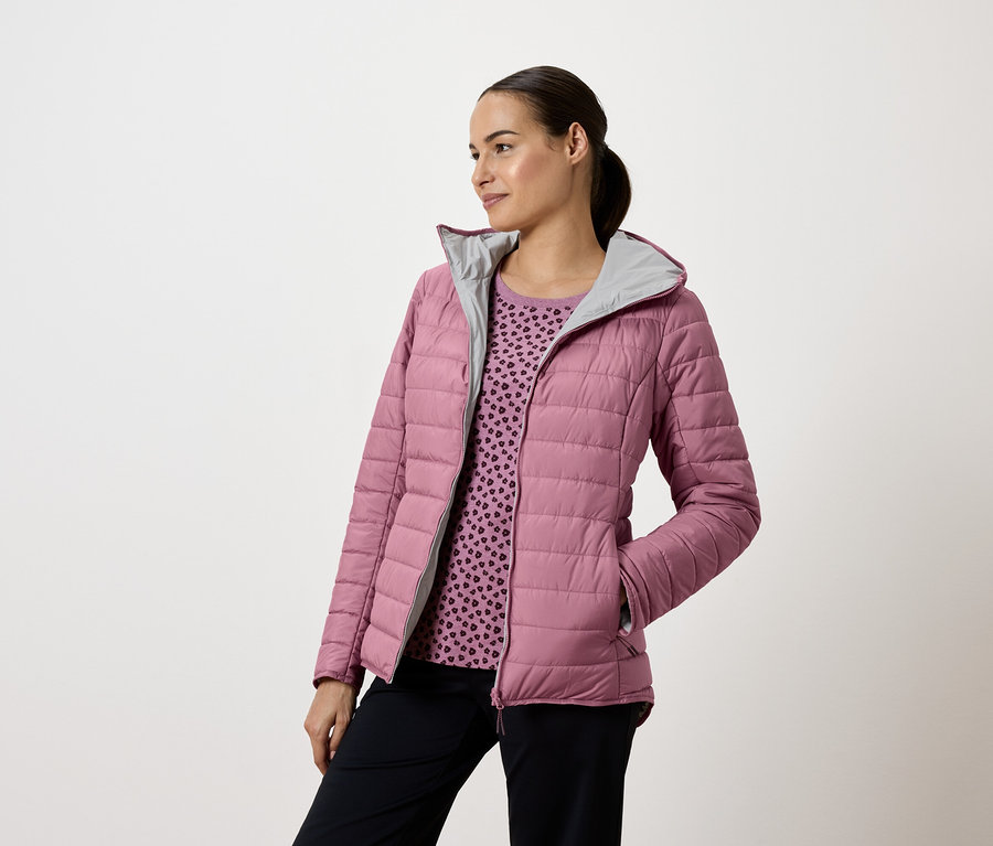 Žena v růžové prošívané bundě s kapucí a tmavomodrých kalhotách, dívá se doleva.