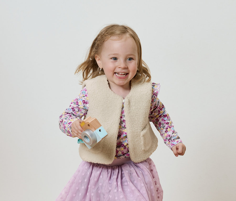 Usmívající se dívka s dřevěným hračkovým fotoaparátem, oblečená v šeříkové halence a sukni, na světlém pozadí.