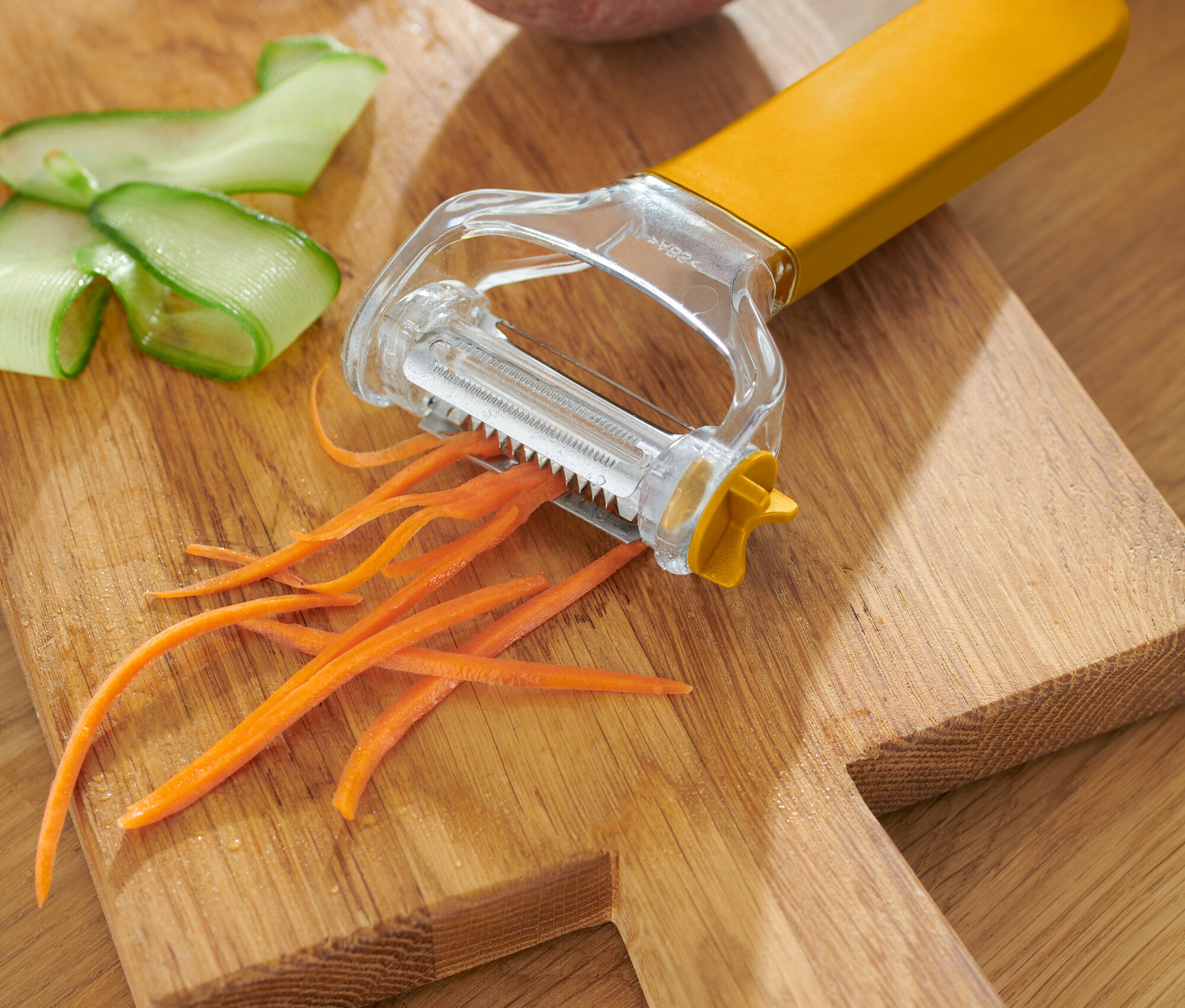 Žlutá škrabka na zeleninu s loupanou mrkví, okurkou a bramborou na dřevěném prkénku.