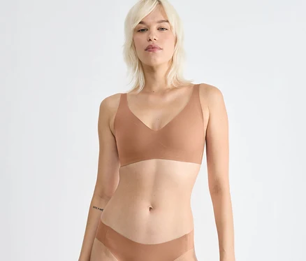 Blondýnka má na sobě měkkou podprsenku sloggi ZERO Feel 2.0 Soft Bra v barvě nostalgic brown a odpovídající spodní prádlo, dívá se dopředu. Má tetování na levém předloktí.