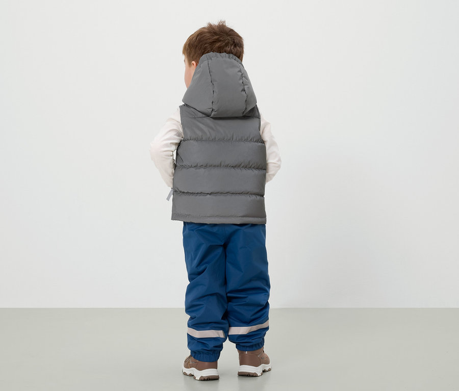 Chlapec stojí zády, má na sobě šedou vestu s kapucí, modré kalhoty a hnědé boty.