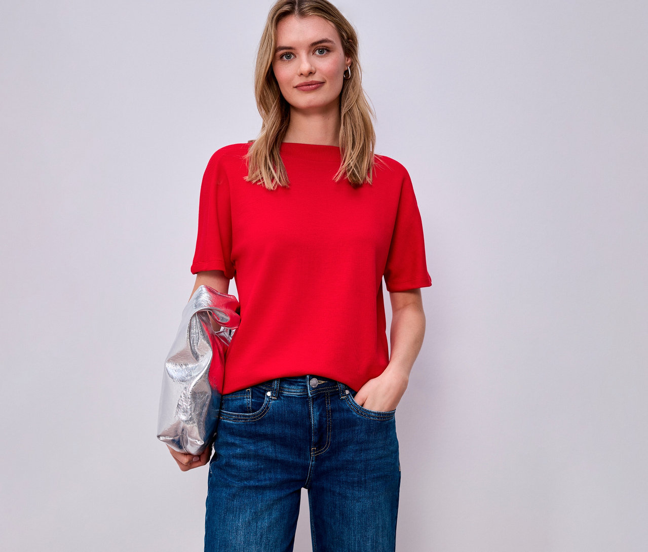 Žena v červeném tričku s krátkým rukávem Street One a džínách, přes ruku má přehozenou stříbrnou bundu.