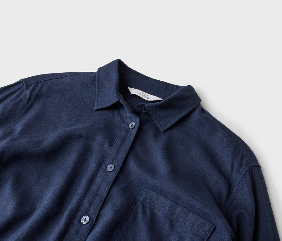 Detailní záběr na tmavě modrou košili s límcem, knoflíky a jednou náprsní kapsou na neutrálním pozadí.