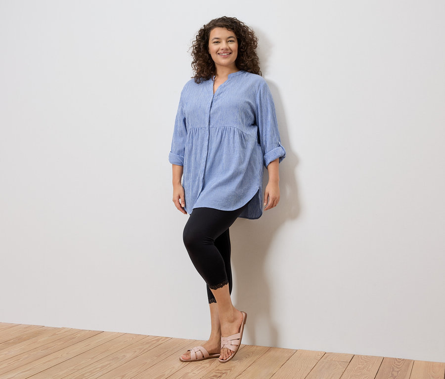 Žena stojí v modré košili, černých legínách s krajkou a růžových sandálech před bílou zdí.