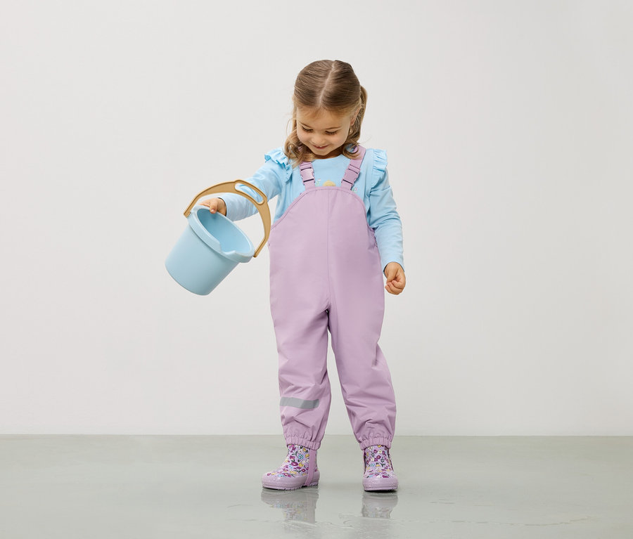 Dívka ve světle modré halence a fialových kalhotách drží světle modrý kbelík.