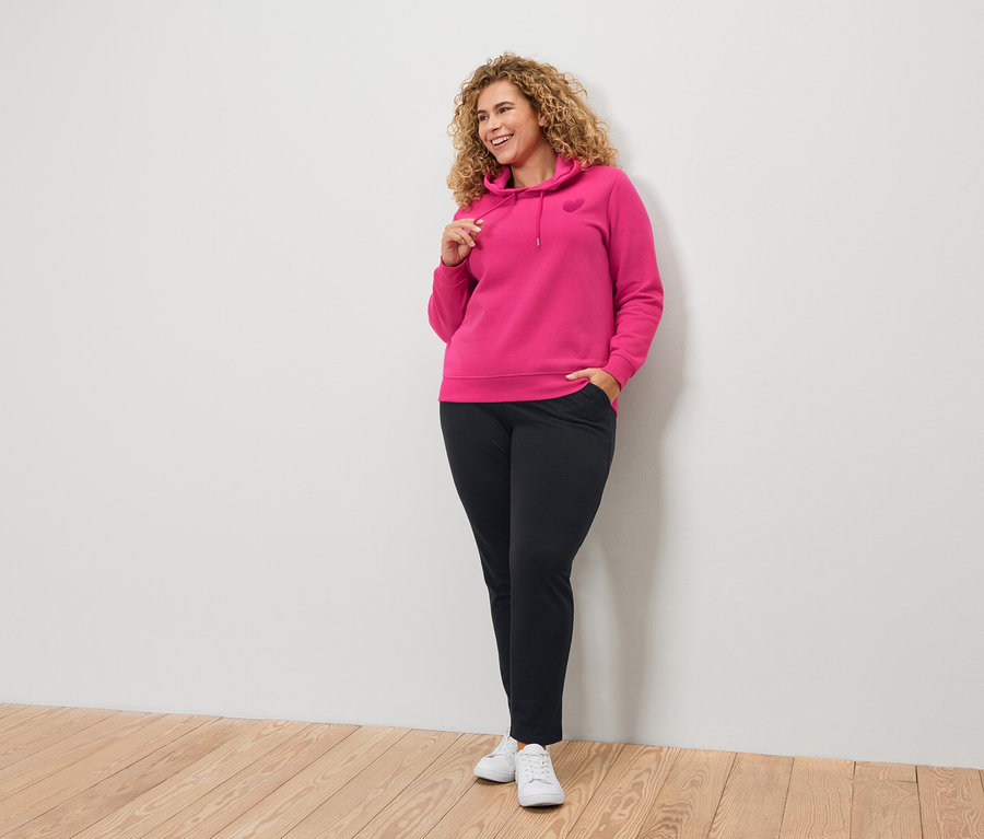 Žena s kudrnatými vlasy v růžové mikině s kapucí a černých kalhotách.