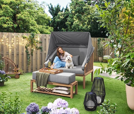 Žena sedí se psem v plážovém koši Premium Lounge-Strandkorb mit Sonnenschutz-Dach. Na trávníku stojí solární lucerna s textilním výpletem, velká a solární lucerna s textilním výpletem, malá. V pozadí je barový vozík s odnímatelným podnosem. Na plotě visí venkovní světelný řetěz s LED a síťovým adaptérem.