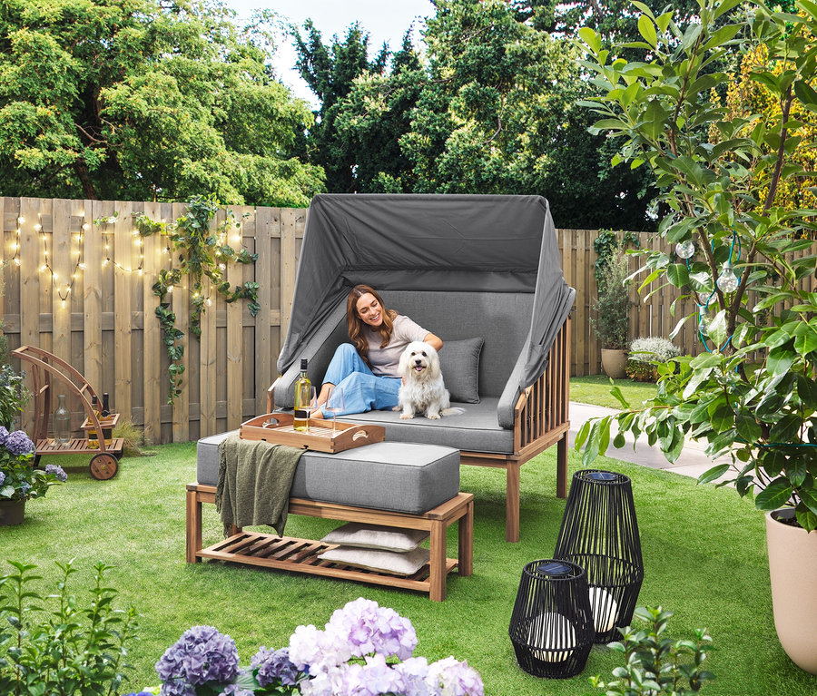 Žena sedí se psem v plážovém koši Premium Lounge-Strandkorb mit Sonnenschutz-Dach. Na trávníku stojí solární lucerna s textilním výpletem, velká a solární lucerna s textilním výpletem, malá. V pozadí je barový vozík s odnímatelným podnosem. Na plotě visí venkovní světelný řetěz s LED a síťovým adaptérem.