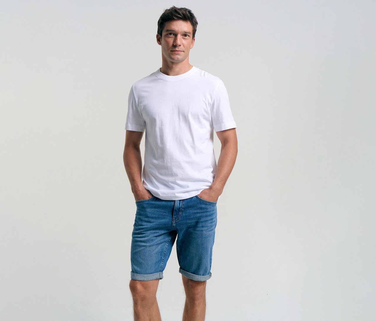 Muž v bílém tričku a džínových šortkách Tom Tailor TTJOSH REGULAR SLIM, mid stone bright blue denim, s rukama v kapsách.