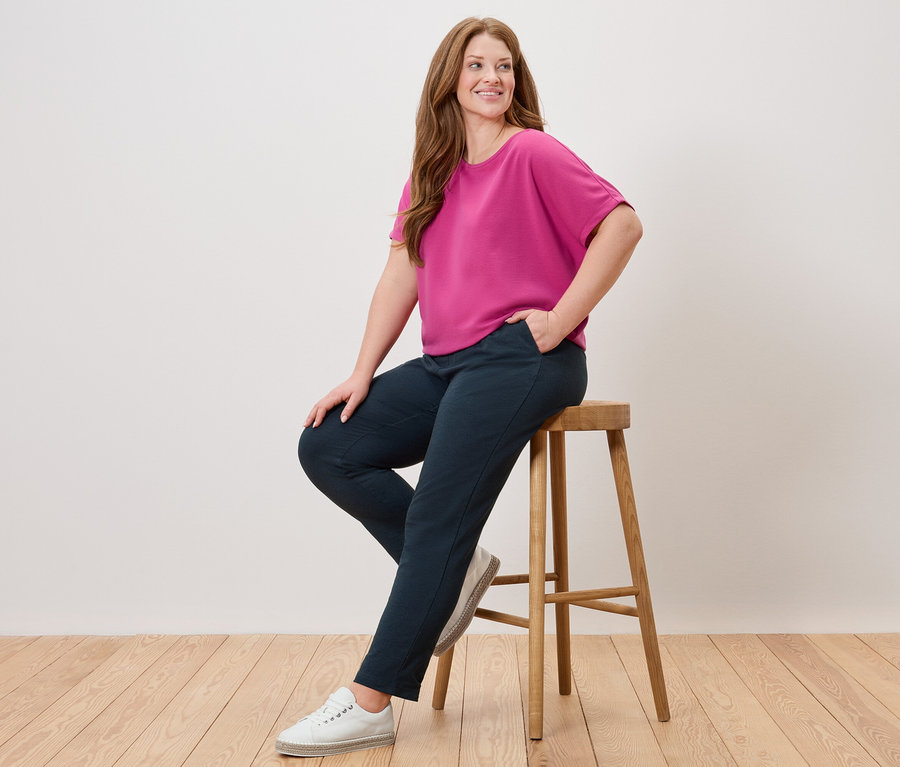 Žena sedí na stoličce, má na sobě růžovou halenku, modré kalhoty a bílé boty.