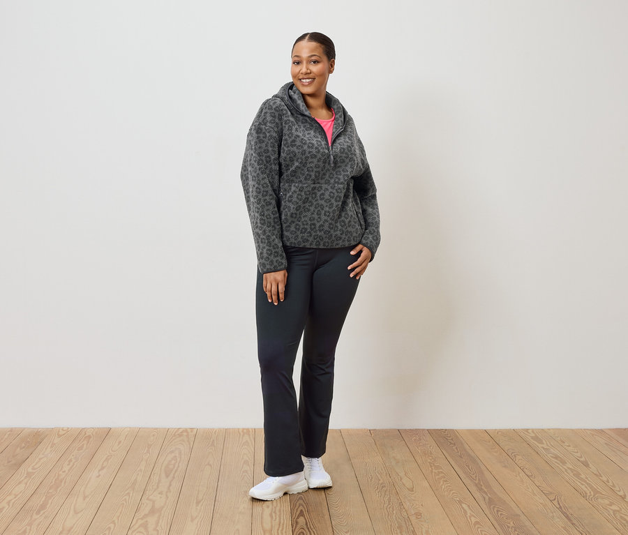Žena stojí a má na sobě šedo-černou leopardí fleecovou mikinu s kapucí, černé kalhoty a bílé tenisky.